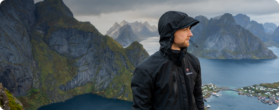 Man in a black heated jacket standing in front of a mountainous landscape with a fjord.